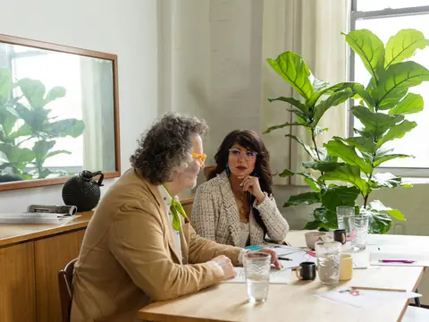Two women talking at a table in a bright room full of plants