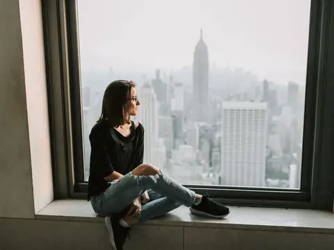 A woman sitting on a wide windowsill looking out at the Manhattan skyline and the Empire State Building
