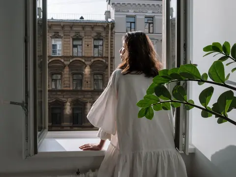 A woman in a white dress at an open window looking out at old city buildings on a sunny day